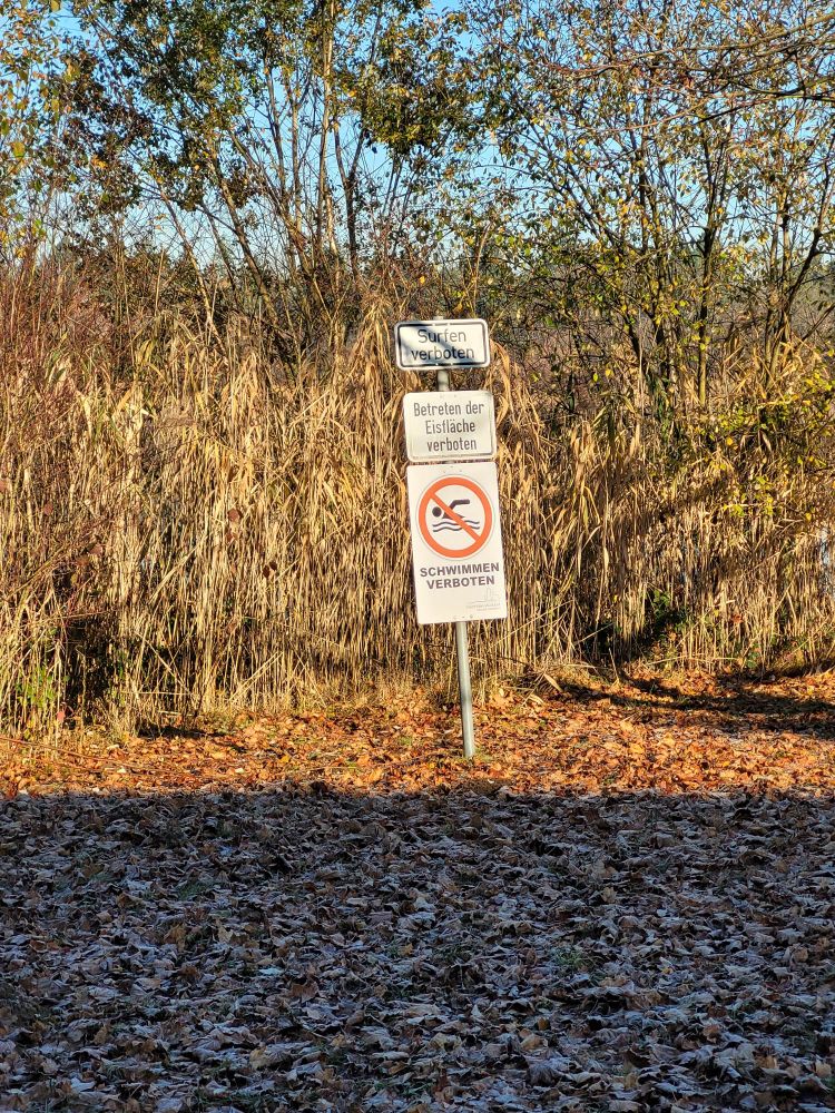 Verbotsschilder an einem Baggersee, von oben nach unten:
Surfen verboten
Betreten der Eisfläche verboten
Schwimmen verboten