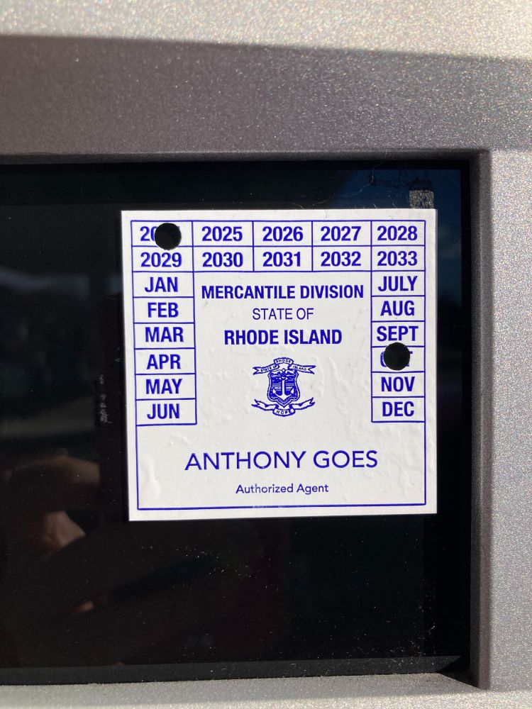 A photo of a sticker with blue text on a white background. The photo has "2024" and "OCT" punched out from a selection of years and months, and features the text "MERCANTILE DIVISION STATE OF RHODE ISLAND" and the name "ANTHONY GOES Authorized Agent"