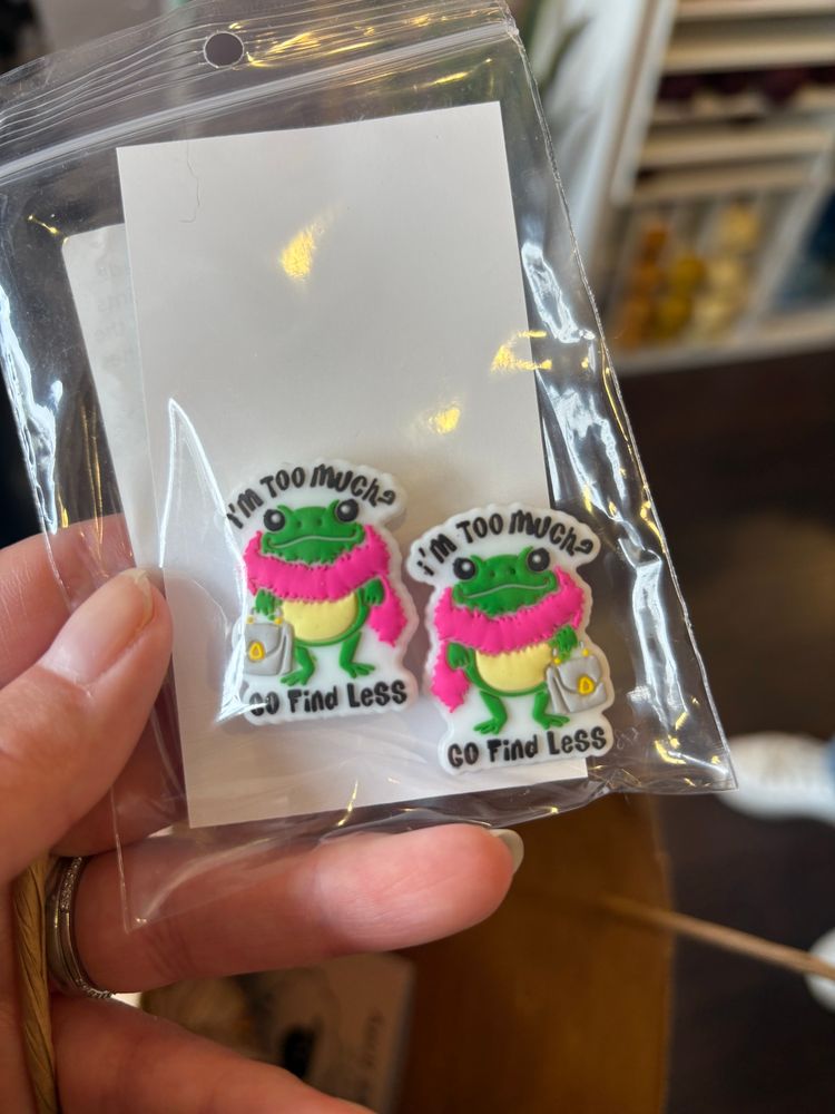 stitch stoppers for knitting needles. They are shaped like a frog wearing a pink boa with the words “I’m too much? go find less” around the frog