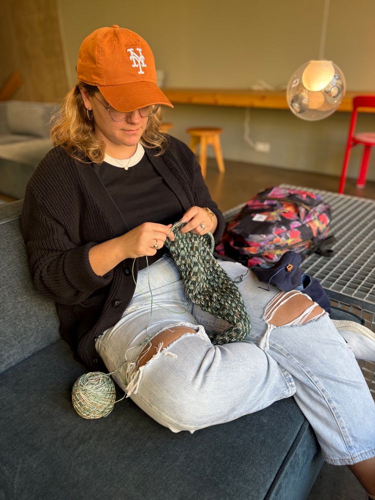 A young woman sits on a couch knitting a scarf, looking down at her project with focus. She is wearing an orange NY Mets hat, a black shirt, and light blue ripped jeans. 
