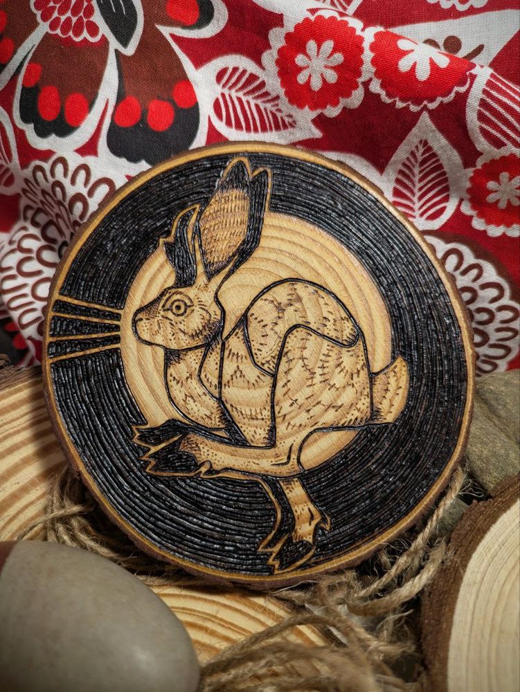 photo of a wood slice with a pronghorn/jackrabbit jackalope burned onto it. the creature is spotlit against a dark bg with a heavy texture of concentric circles. the piece is propped on a red, floral patterned fabric and surrounded by other pieces of wood and stone and jute twine.