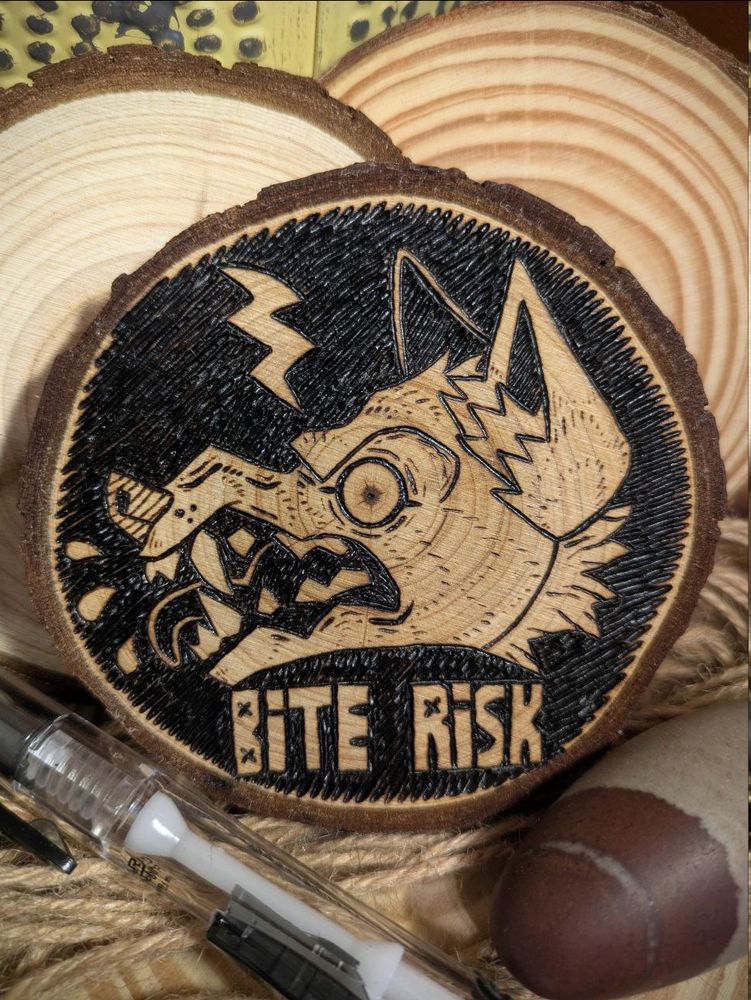 photo of a piece of woodburning art featuring a snarling canine head on a black bg. there is a lightning bold at the top of the piece, as if striking the dog or coming from it, and there are three drops of blood or spit coming from the dog's teeth. the words "BITE RISK" are below the head. the entire piece is on a ~3.5" smooth round slice of wood.