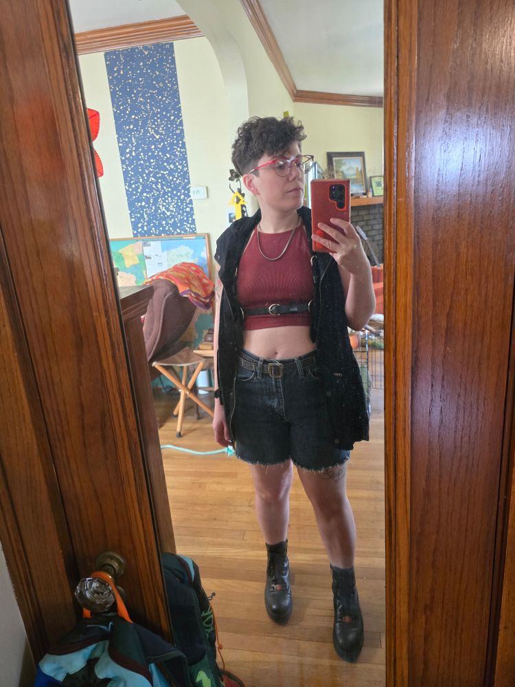 Me but hot af.
AV, an enby standing in a mirror with short brown hair, glasses, mahogany tank top, cut off jorts, leather boots, a silver chain, leather harness, and long black vest.