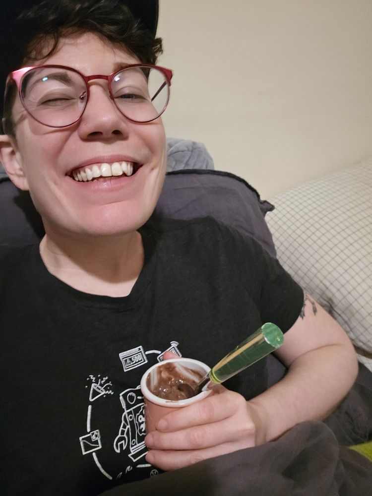 AV, a nonbinary person with short brown hair, a cap, and a black shirt with a stupidly large grin is sitting in bed holding a cup of homemade chocolate pudding with a spoon sticking out of it