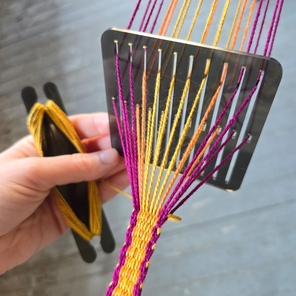A plastic rigid heddle backstrap loom and shuttle with purple, yellow, and orange string. A band is partially finished being woven.