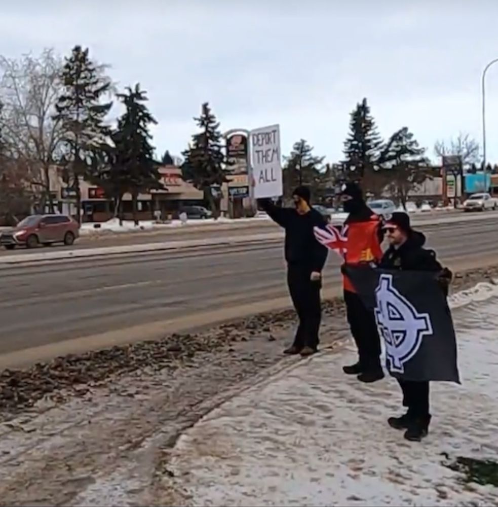 The three men are now? In the same position on the side of the road, holding three separate items. The first has a sign that reads “deport them all,” the second has a red ensign flag (which has been adopted in canada by white supremacists as a symbol for a white canada), and the third is holding a glad with a Celtic cross (another adopted hate symbol used by white supremacists)