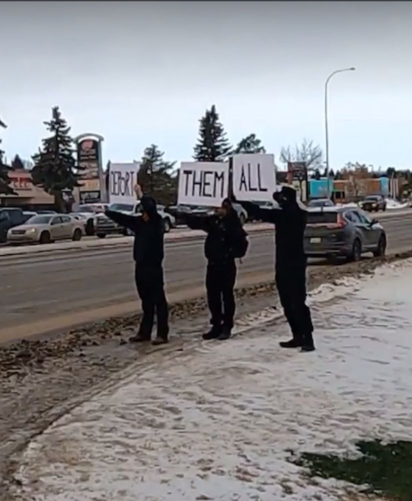 Three men join the first on the side of the road, all in black, each holding a sign that together read “deport them all.” They’re all throwing up Nazi salutes