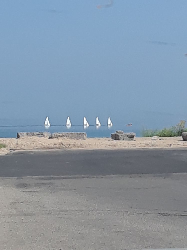 4 small sailboats with white sails are trailing a teaching boat/dinghy in Lake Huron.  The horizon is hardly visible as the sky and water are the same colour.