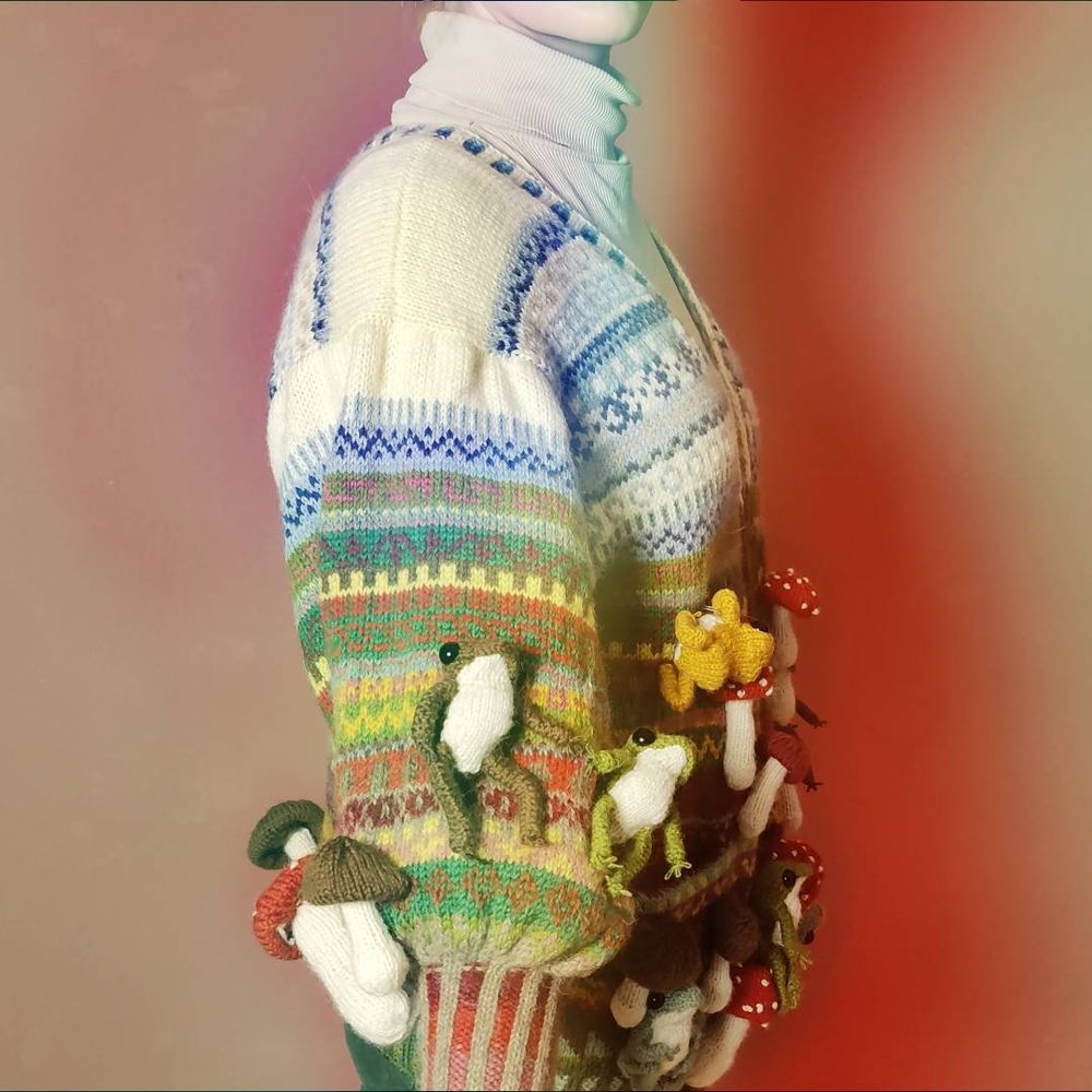 Person wearing a colourful Fair Isle style cardigan with many knitted frogs and mushrooms attached all over