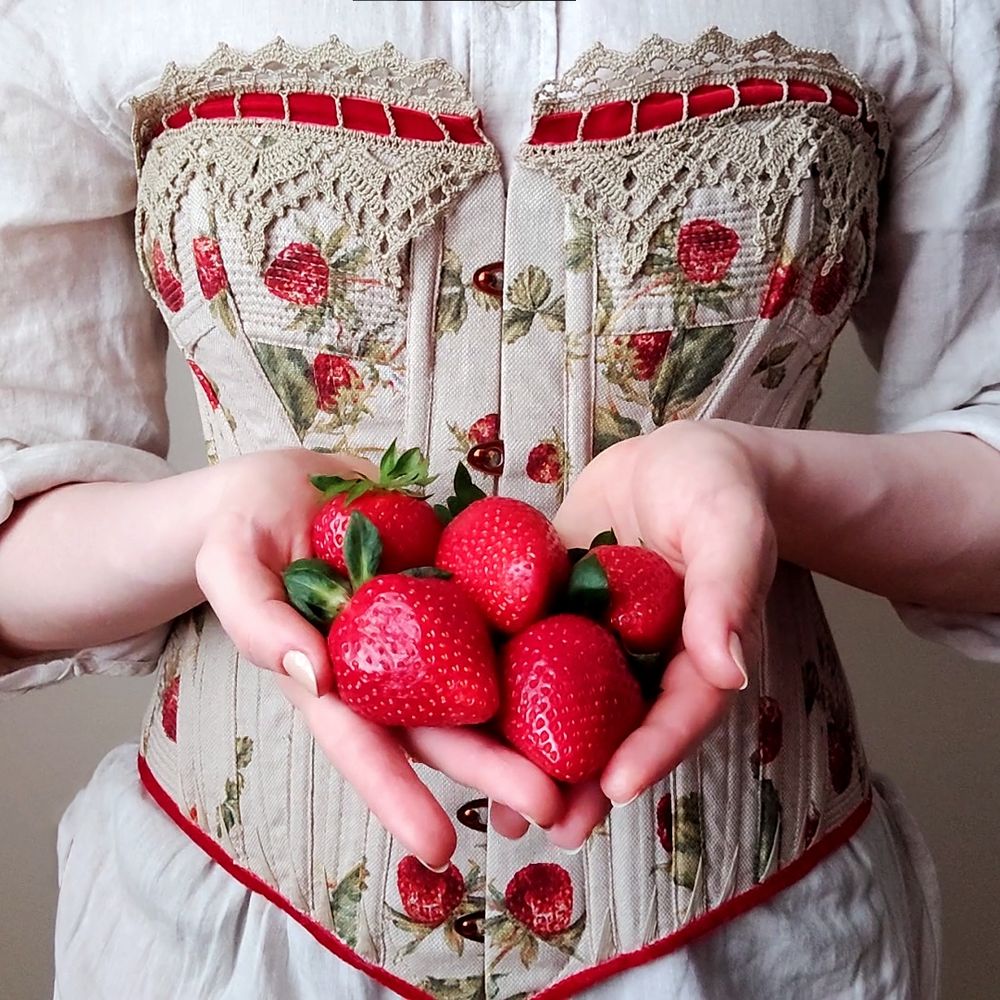 late 19th century style corset made in beige fabric with strawberry print. top edge of corset has beige crocheted lace with a red velvet ribbon through it. model is holding a handful of strawberries.