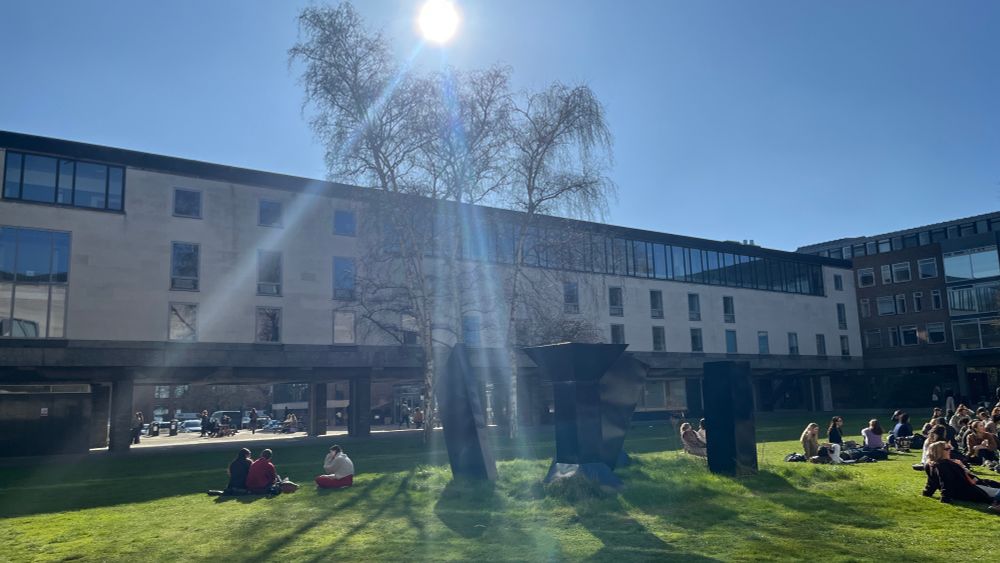 The grass under the Raised Faculty Building on a sunny March morning, with groups of students sat around talking.