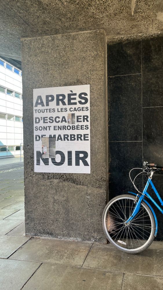 On a concrete pillar of the Raised Faculty Building, a poster in French reads “Après toutes les cages d’escalier sont enrobées de marbre noir. Smaller posters in English translation are superimposed on the French one.