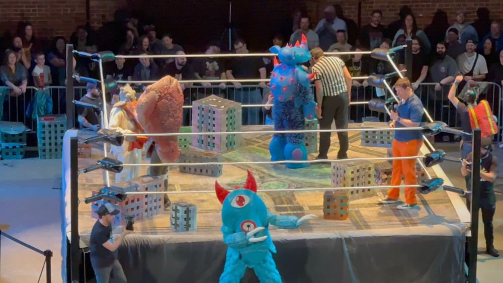 A picture of a wrestling match with four kaiju fighting, plus a crowd in the background and staff seen as well. 