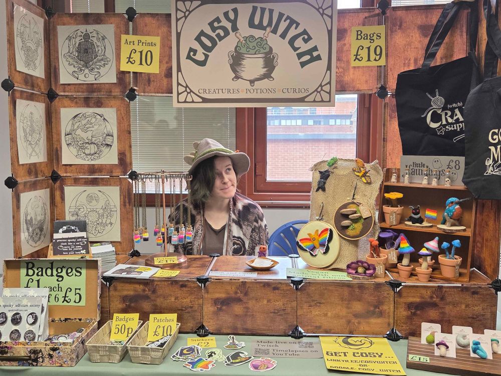 A magical market stall with a big sign with a cauldron and the words Cosy Witch on it.the stall is covered with needle felted goodies, witchy prints and badges.