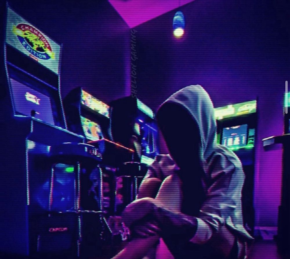 A gamer woman with a tattoo sleeve on her left arm. She wears a yellow hoodie, sitting quietly in a dimly lit arcade game room, highlighted by the game rooms neon lighting.