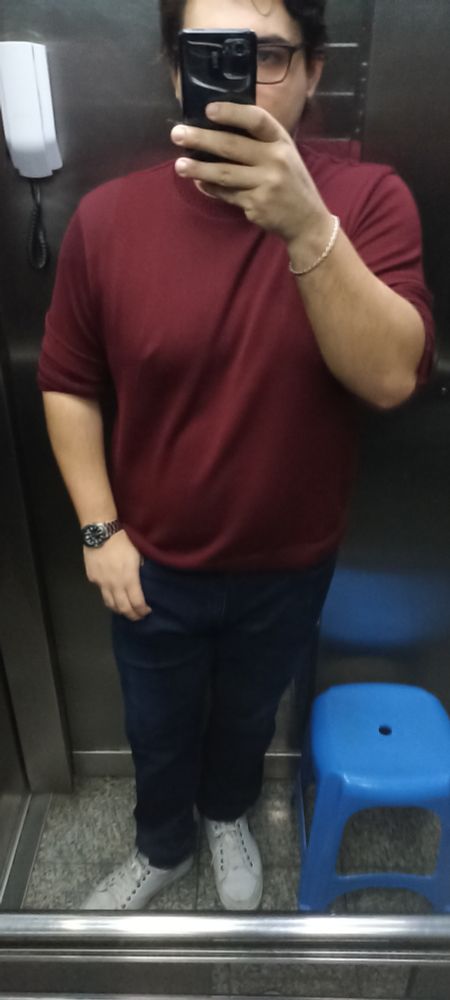 "selfie" em pé no elevador. Estou com um suéter vinho, jeans azul e tênis branco. Há uma banqueta azul do meu lado.