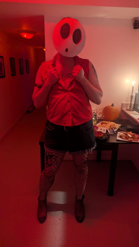 Owen with the same costume on the bottom half and the mask, with a red shirt with the sleeves ripped off, holding the lapels of the open shirt. 