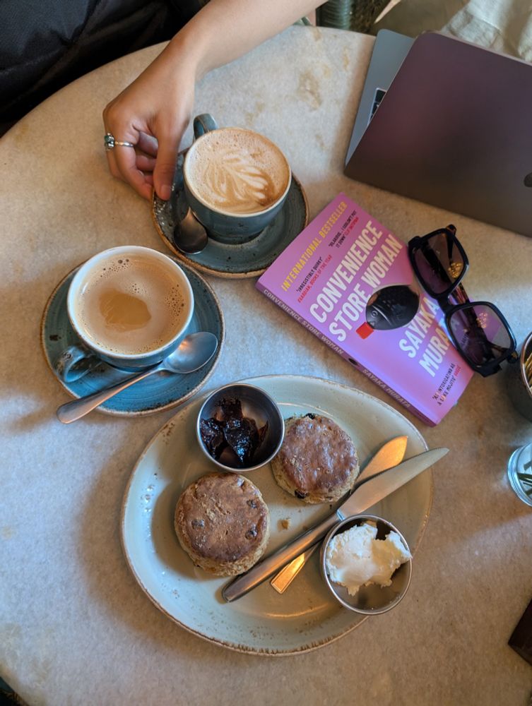 A table at a cafe with a plate of scones, a cup each of coffee and chai and a book with sunglasses on top of it, with a laptop nearby.