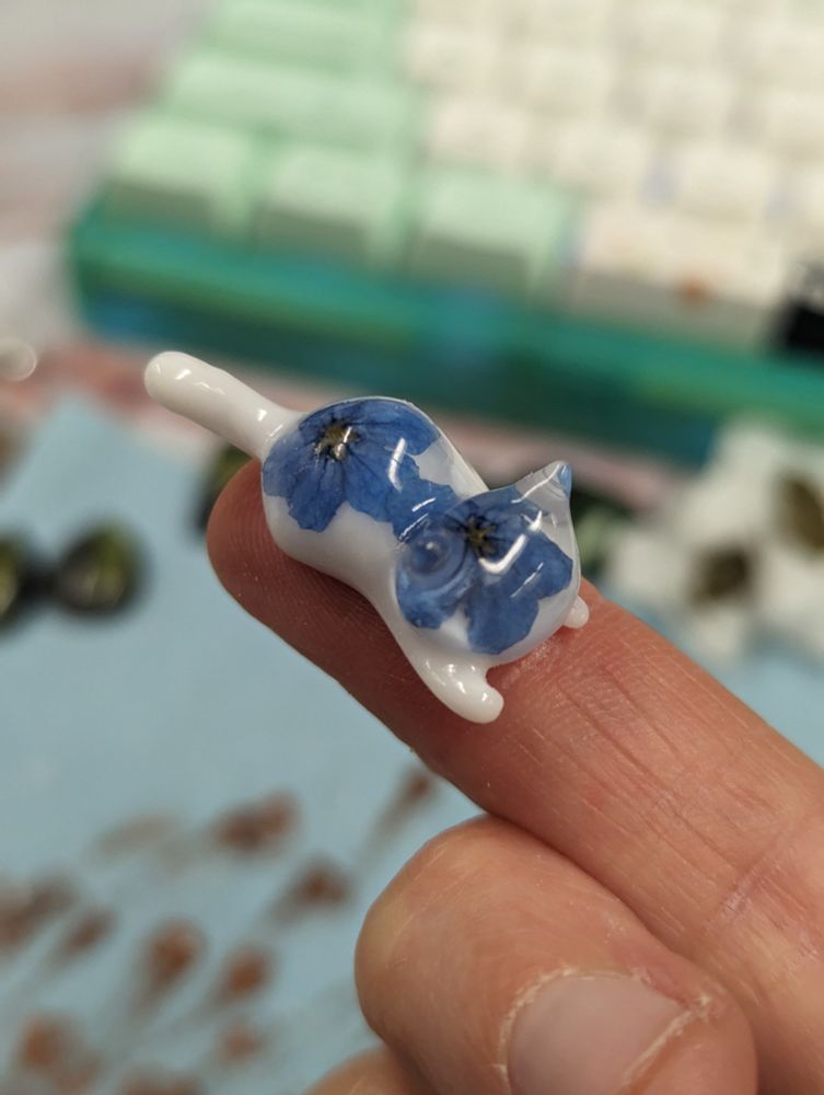 A small resin cat with forget me not flowers in it resting on top of an outstretched index finger