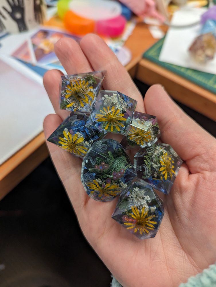 An uninked 7-piece polyhedral dice set held in a hand and made using alternate shapes with real flowers and a small yellow flower shown in the middle of each die.