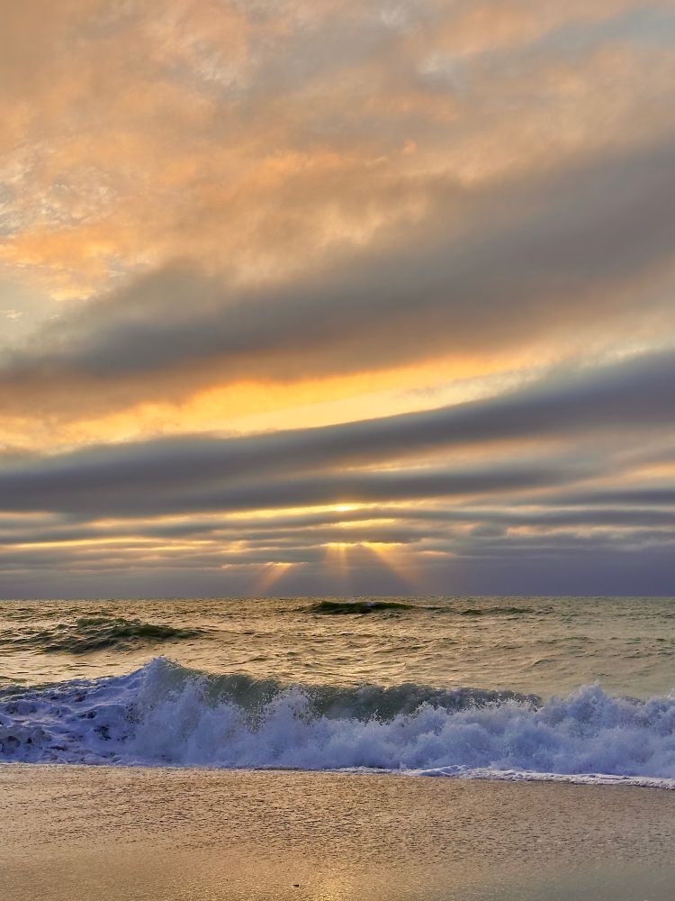 Streaks of dark clouds in front of the setting sun. Rough waters with waves. Sun reflection on the beach sand.