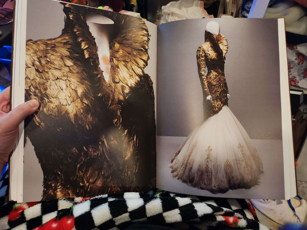 A photo of a book showing 2 views of an Alexander McQueen dress shaped near identically to Agatha's artwork dress. Black and gold feathers cover the upturned collar, long sleeves, and down to the knees, where white chiffon billows out.