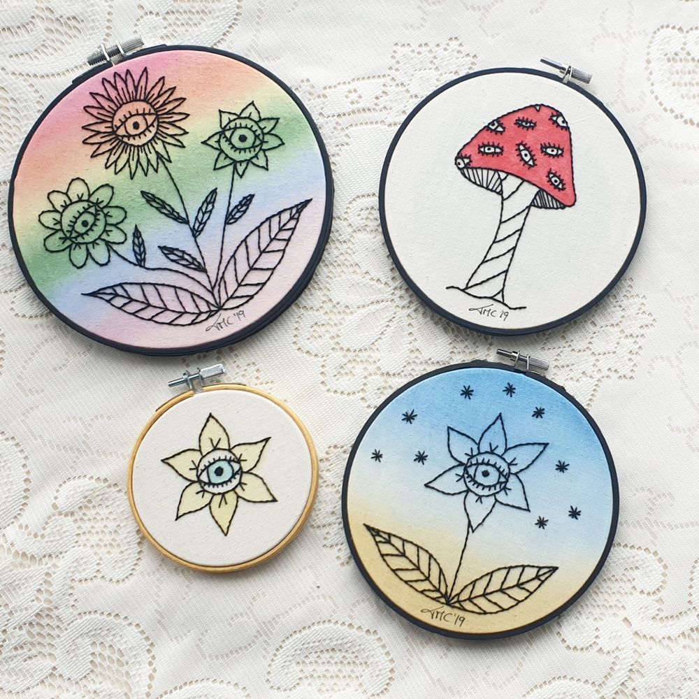 Four embroidery hoops lying on a cream lace table cloth. The hoops have been set with light coloured fabric and a variation of flowers with eyes at the centre of their heads have been stitched in black thread and then painted with fabric paints or markers.