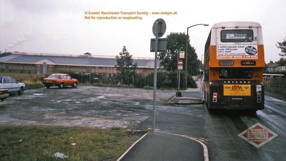 A 1980s view shows a bus on the right, heading away from us. On the left there are a few cars parked on some waste ground and beyond that, a large building that's long and low. It's made from brick with a sloping slate roof.