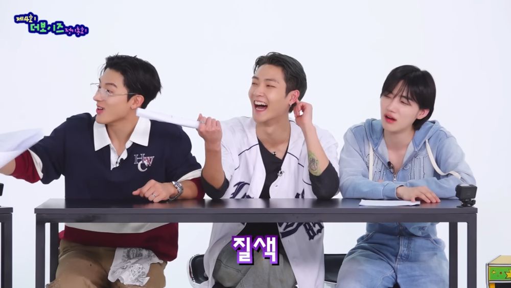 THE BOYZ Eric, Kevin, and New (Chanhee) sitting together. They're all looking to Hyunjae who's out of frame, and while Eric and Kevin are smiling brightly, Chanhee is making a face and in the middle of saying "I hate it".