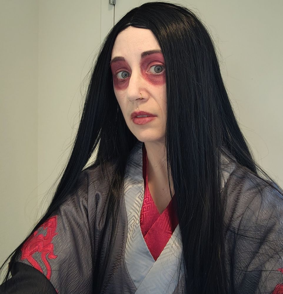 A shoulders-up photograph of a person dressed as Mo Xuanyu from Mo Dao Zu Shi. They are wearing dark and light grey robes with red trim, a long black wig, grey contact lenses, and white face makeup with red lips and red around the eyes. Their expression is sad and a little scared.