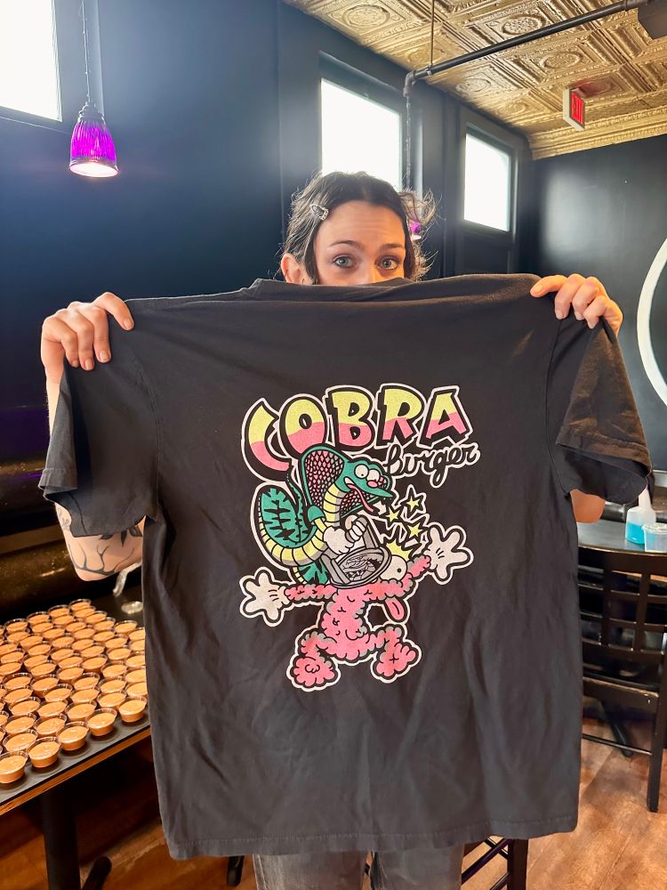 Oliv, the bar manager at Cobra Burger holding the a T-shirt. On the shirt is a cartoon cobra smashing a cartoon beef patty monster