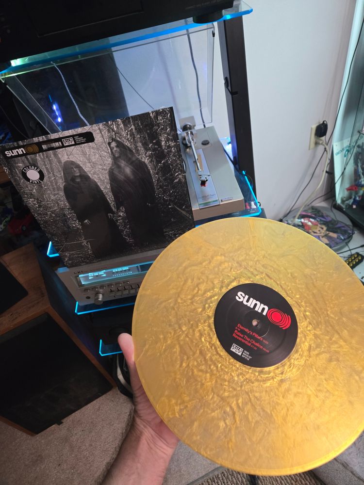 The gold sparkling colored record for the band sunn0))) with the album art in the background featuring the two band members in their typical cloaks in black and white in a forest.