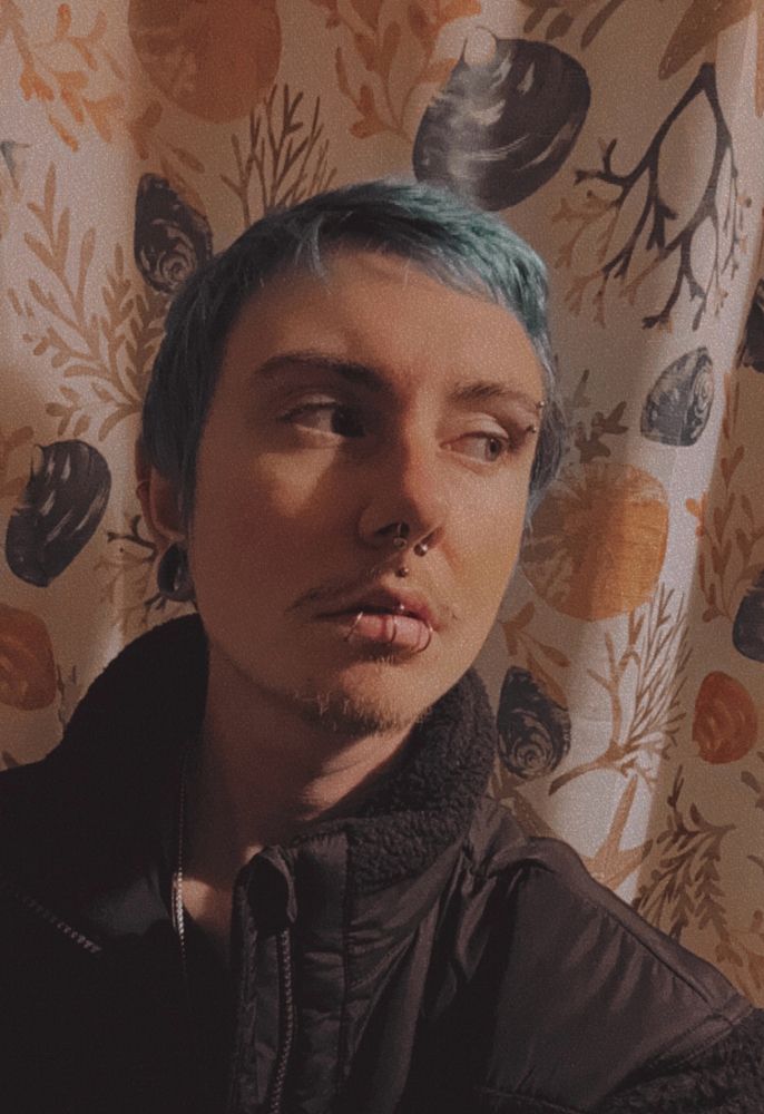 A man with blue hair and facial piercings (eyebrow, nose ring, four lip piercings, stretched ears) wearing a black coat and looking off into the distance. 