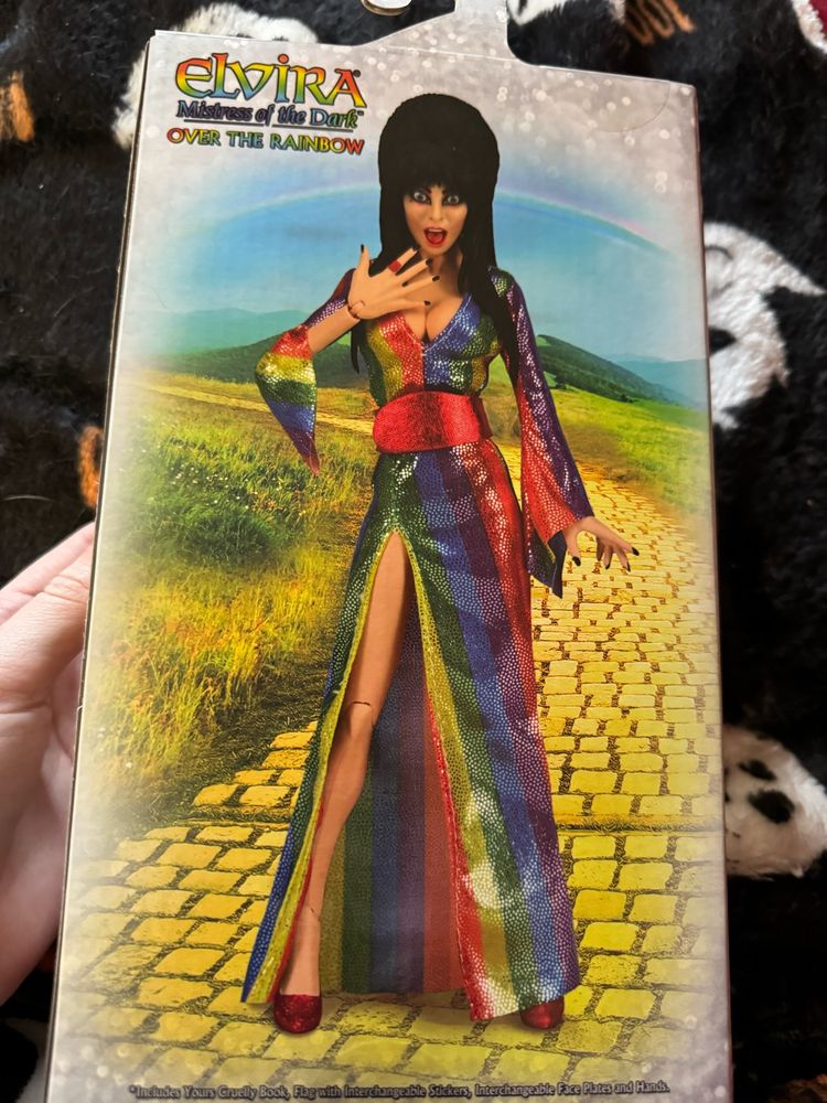 Photo of a posable Elvira action figure wearing a rainbow dress. Elvira is standing on a yellow brick road with a surprised expression on her face. 