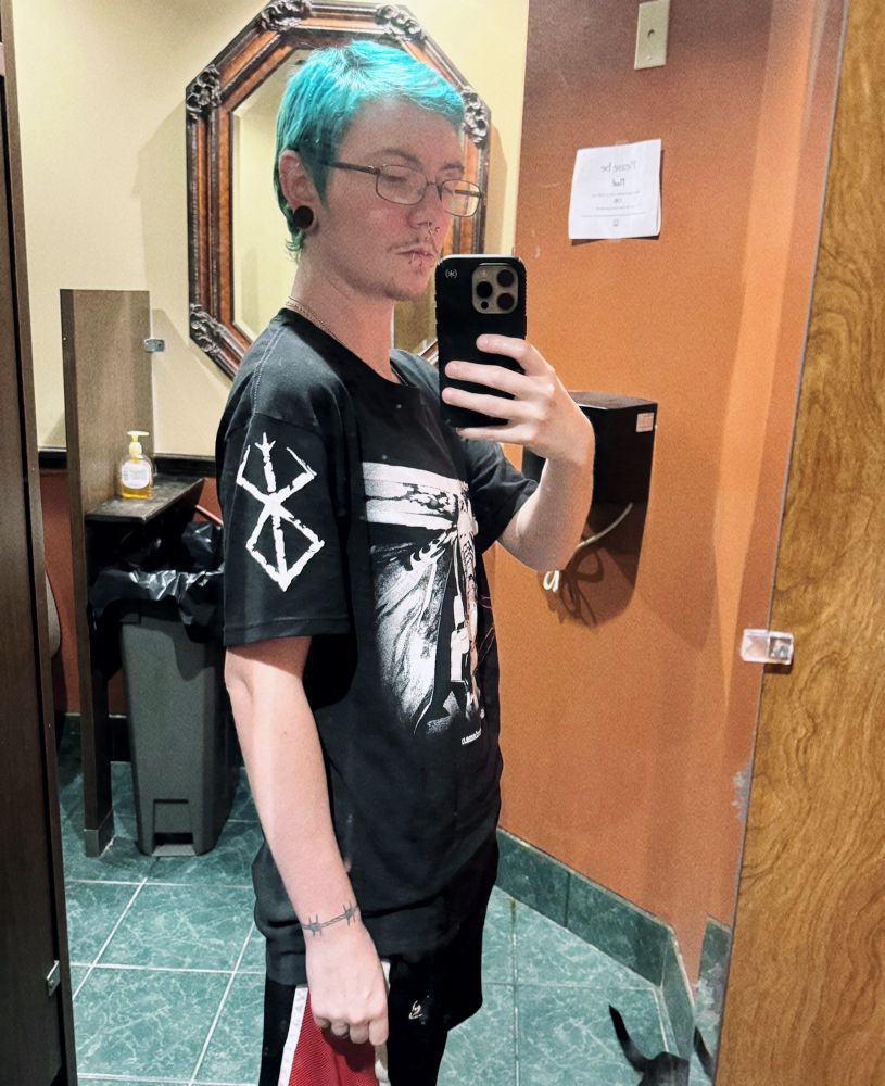 A thin, white man with dyed blue hair posing in front of a restroom mirror. He is standing with his side profile facing the mirror, showing off the sleeve design on his black graphic t-shirt. There is a cryptic symbol on the left sleeve from the manga "Berserk." He is wearing glasses and has multiple facial piercings as well as stretched ear lobes. In the photo, he has a wrist tattoo of barbed wire on his left wrist. 