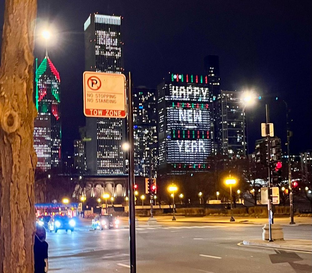 An evening city scene. A lone tree on the left foreground next to a no parking signs. Tall, lit buildings in the background. One of the buildings chose to program their lights to spell “Happy New Year.” 