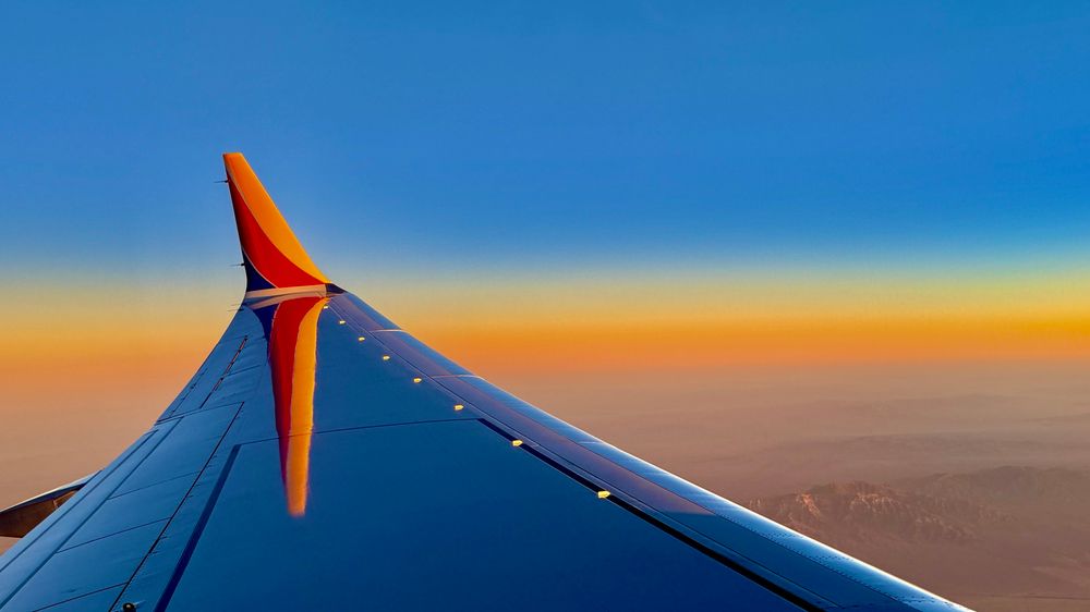 This photo shows the wing of an airplane in flight, during sunset. Here's a breakdown of the key elements:
 * Foreground: The wing of the airplane dominates the foreground, with a prominent display of the airline's livery (the color scheme and logo). The wing's surface reflects the colors of the sky, creating an interplay of light and shadow.
 * Background: The background showcases a vibrant sunset with a mix of orange, yellow, and blue hues. The landscape below is a mountainous desert region, flying over the southwestern United States.
 * Composition: The photo's composition uses the lines of the wing to lead the viewer's eye towards the sunset, creating a sense of depth and movement. The contrast between the cool blue tones of the wing and the warm colors of the sunset adds visual interest.
 * Mood: The overall mood of the photo is one of serenity and awe, capturing the beauty of air travel and the vastness of the sky. The sunset adds a touch of drama and romance to the scene.

The airplane is a Southwest Airlines Boeing 737.
