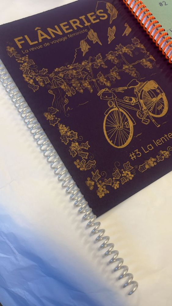 Couverture du prochain Flâneries sur le thème de la lenteur.
Papier violet, encre dorée, spirale nacrée.
La couverture représente un vélo au milieu des vignes avec une montagne au fond. 