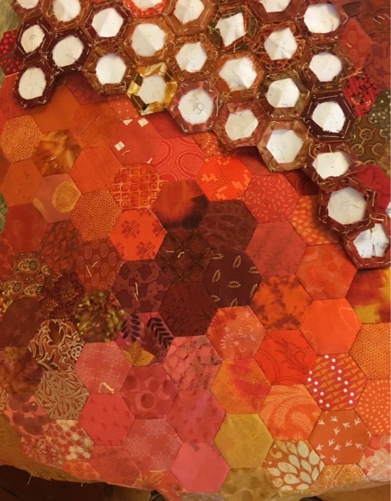 Small hexagons of mainly orange, brown and gold fabric (right side up) being sewn to new larger section that is mainly red and brown (back of project seen).