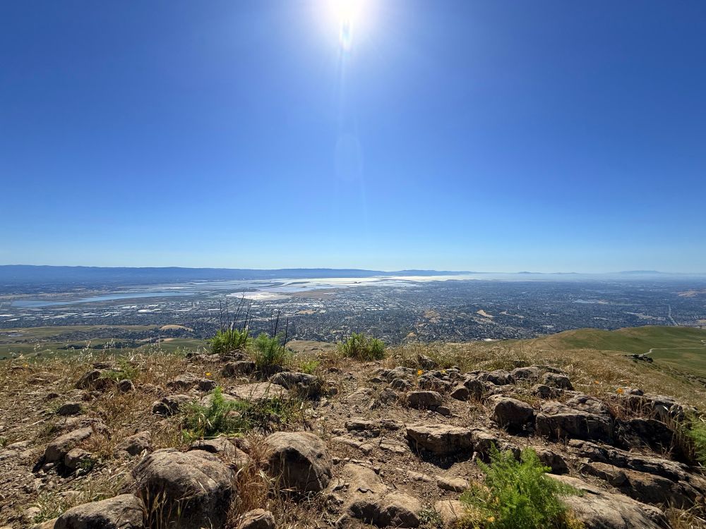A wide angle view of the Bay Area from Mission Peak. In the foreground are gray stone and scrub, the sun hangs over the top center frame. The bay is shimmering.