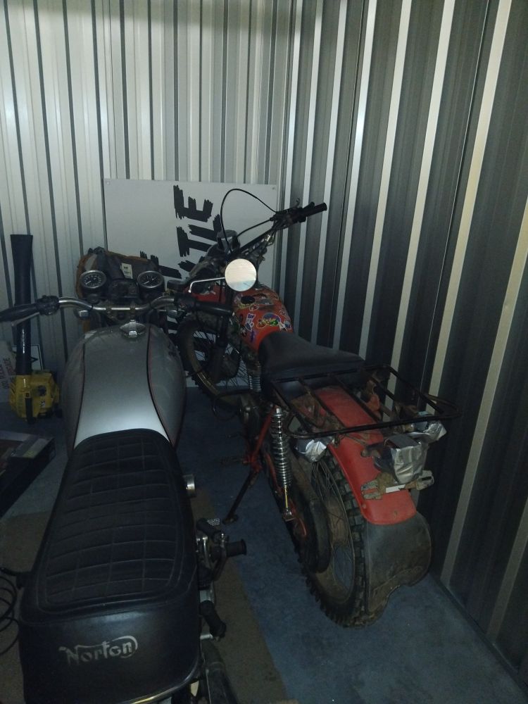 Two motorcyles, a Honda CT125 Trail and a Norton Commando 850, next to each other in a storage shed. The Hondas petrol tank is covered in furry stickers.