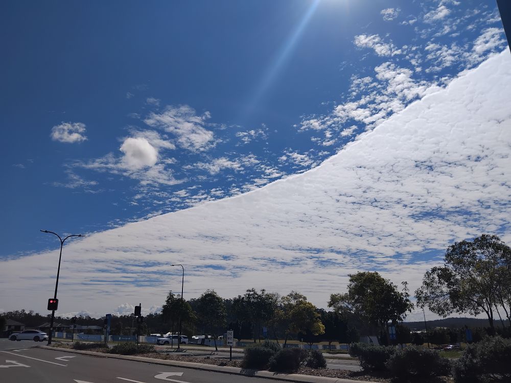 Clouds in an interesting formation over a road intersection. They form a continuous line across the image, diving the sky into a side with almost total coverage and one where the sky is clear and blue, save for a few outliers and patches. 