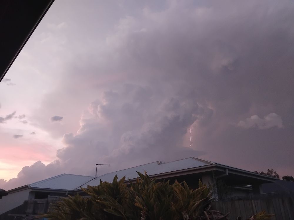 Above the roof of a suburban home towering purple cumulonimbus clouds loom. A single lightning bolt arcs downward between the clouds. 