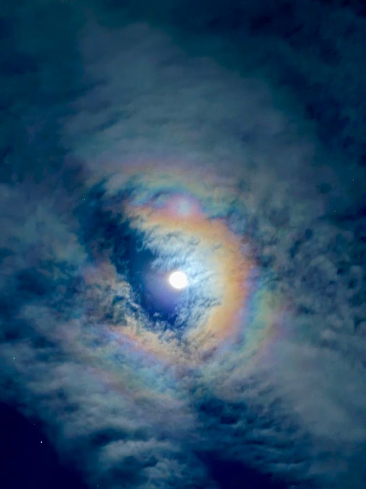 The moon surrounded by a vivid, colorful circular halo of refracted light. The clouds appear thicker and textured around the halo.
