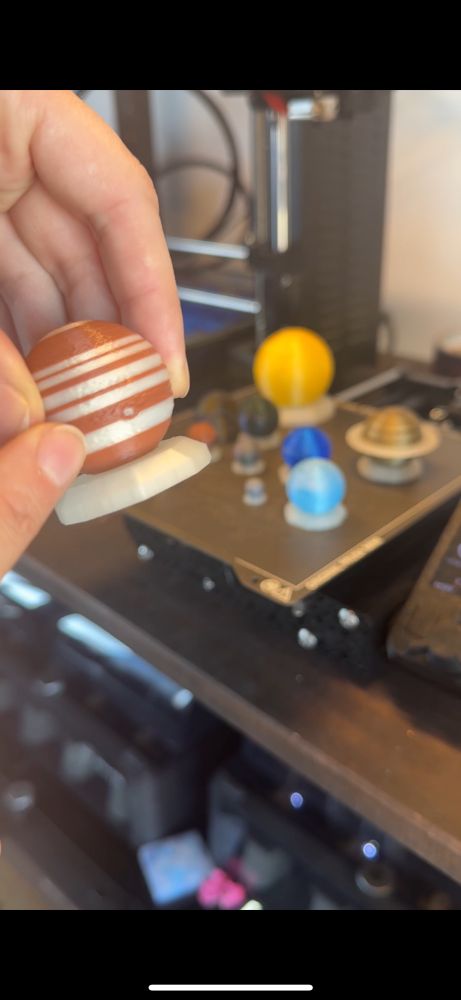 A hand holding a 3D-printed miniature model of Jupiter with brown and white stripes. In the background, there is a 3D printer and other planetary models, including Saturn and Earth, placed on a workbench.