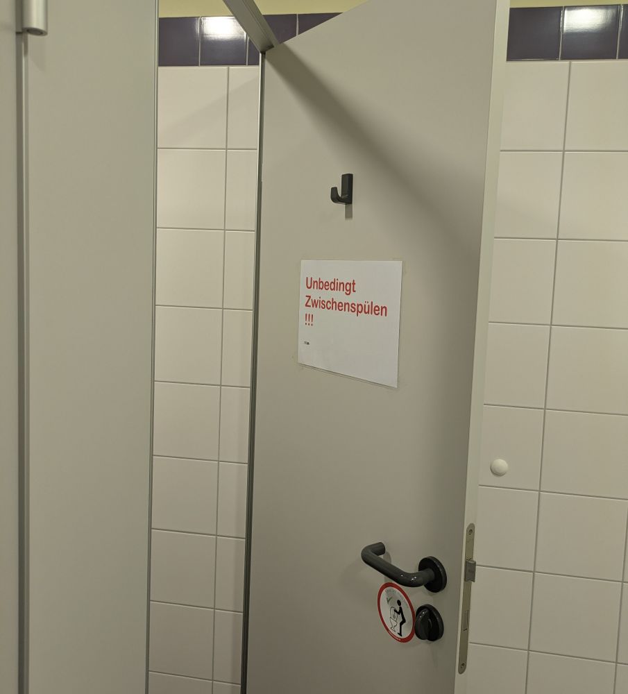 Eine geöffnete weiße Tür einer Klokabine. Auf einem Zettel an der Innenseite der Tür steht "Unbedingt Zwischenspülen!!!" in roter Schrift. Unterhalb der Türklinke ist ein Aufkleber mit einem Piktogramm, das darauf hinweist eine Klobürste zu benutzen. 