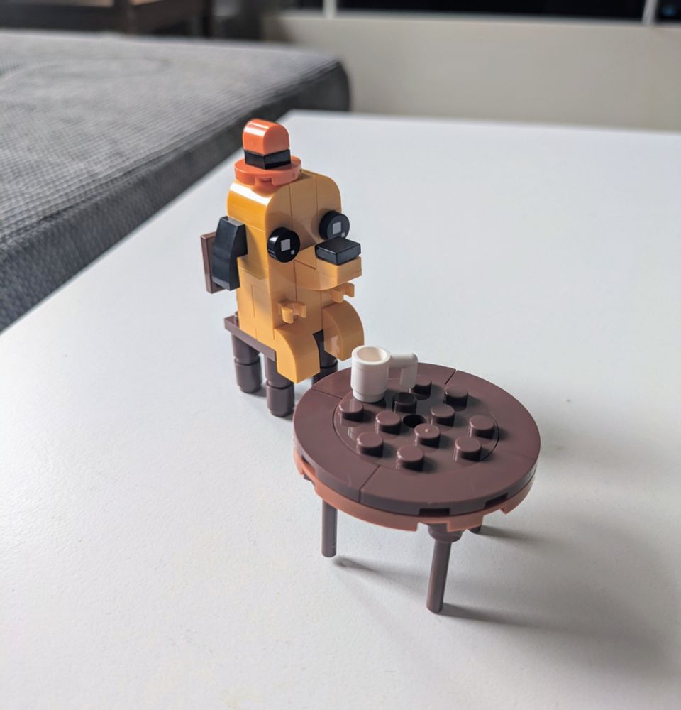 Das Meme "This is fine" aus Legosteinen nachgebaut. Ein orangener Hund mit Hut sitzt auf einem Stuhl an einem Tisch mit einer Tasse darauf. 