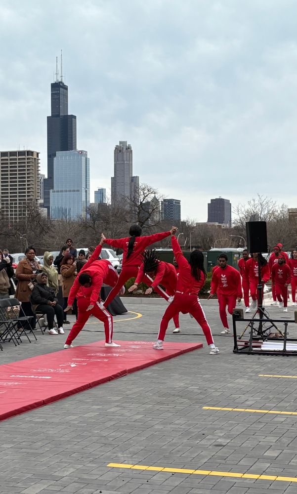 Jesse White Tumblers dressed in red—2 tumblers diving through the arms of three tumblers stacked in a pyramid with the city skyline in the background