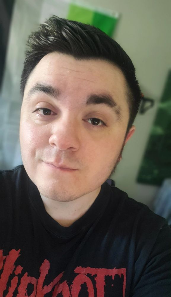 Selfie of RogueDanleo! He has short, dark hair and is wearing a black t-shirt with red lettering. His expression is gentle, and is looking straight at the camera. The background is a bit blurry, but it looks like a room with a green and white flag on the wall.