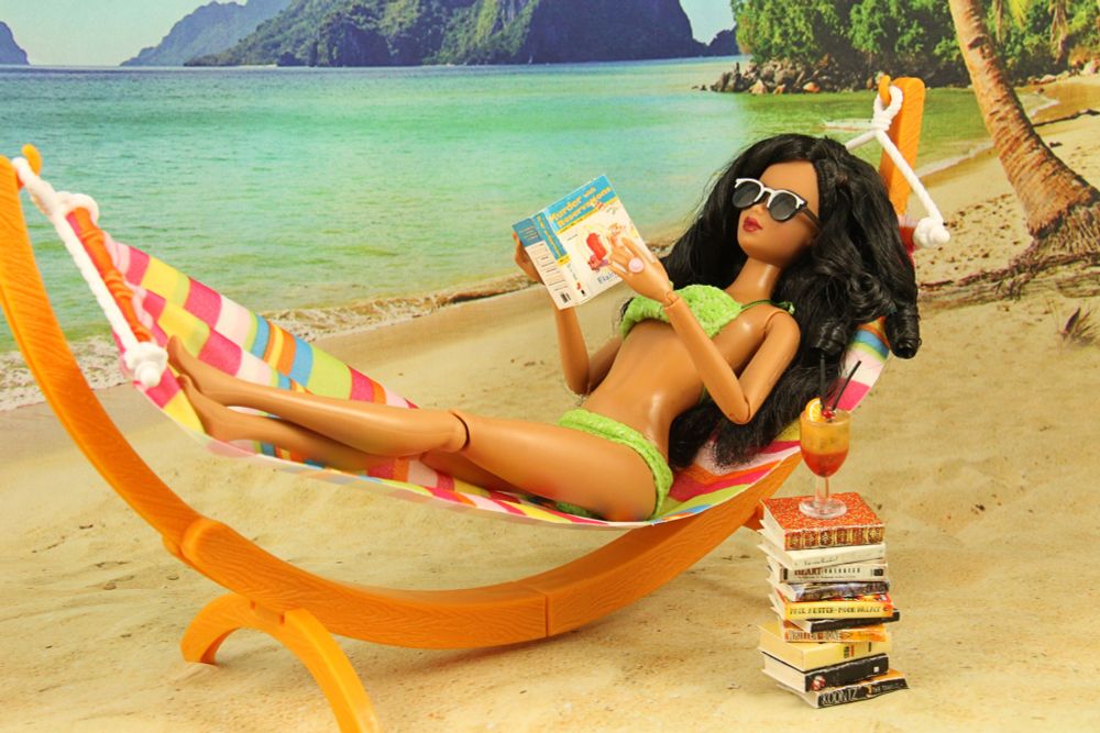 Barbie doll in a green bikini reading books while laying in a hammock on the beach.